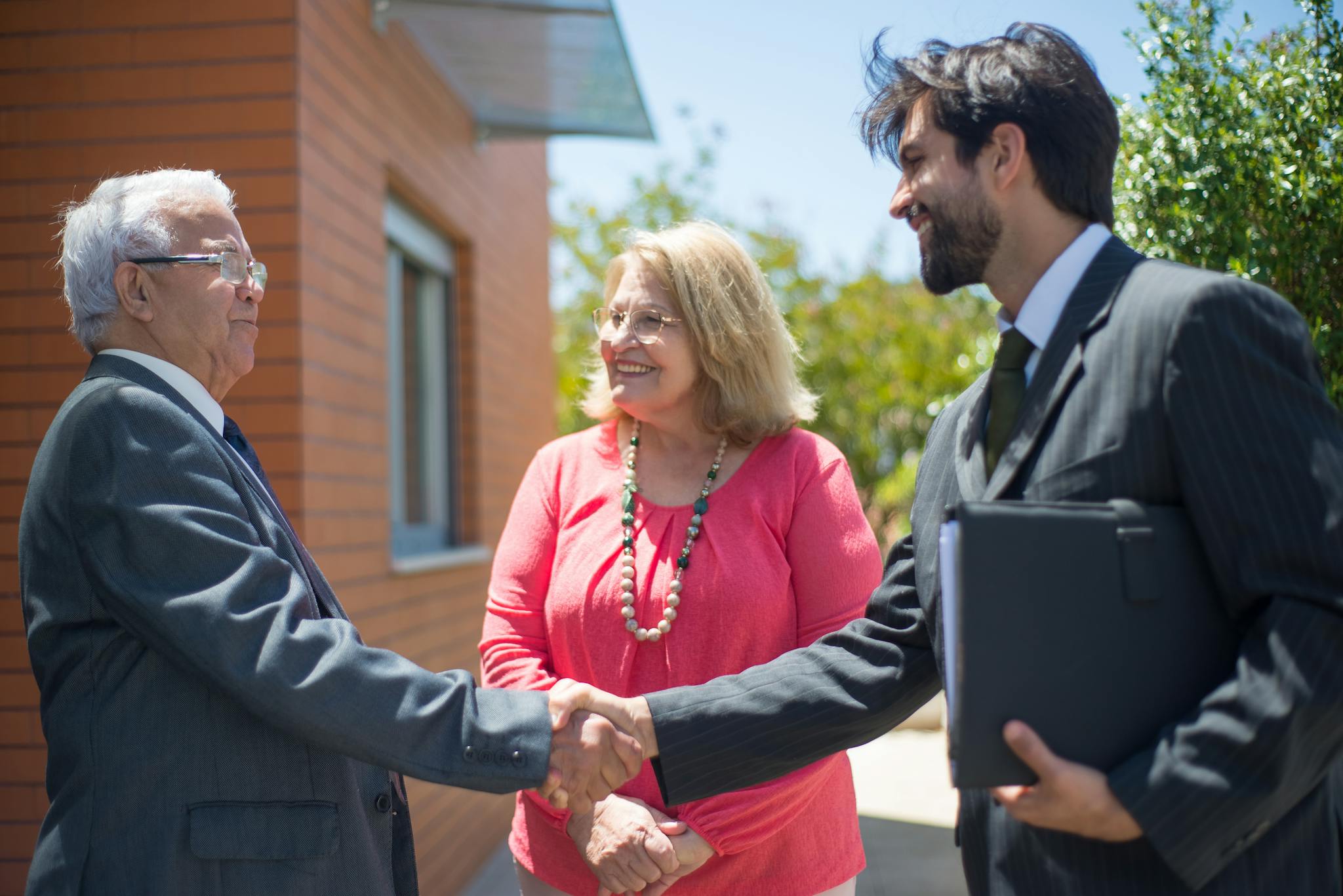 Real estate agent and mature couple shaking hands outdoors, sealing a deal with smiles.